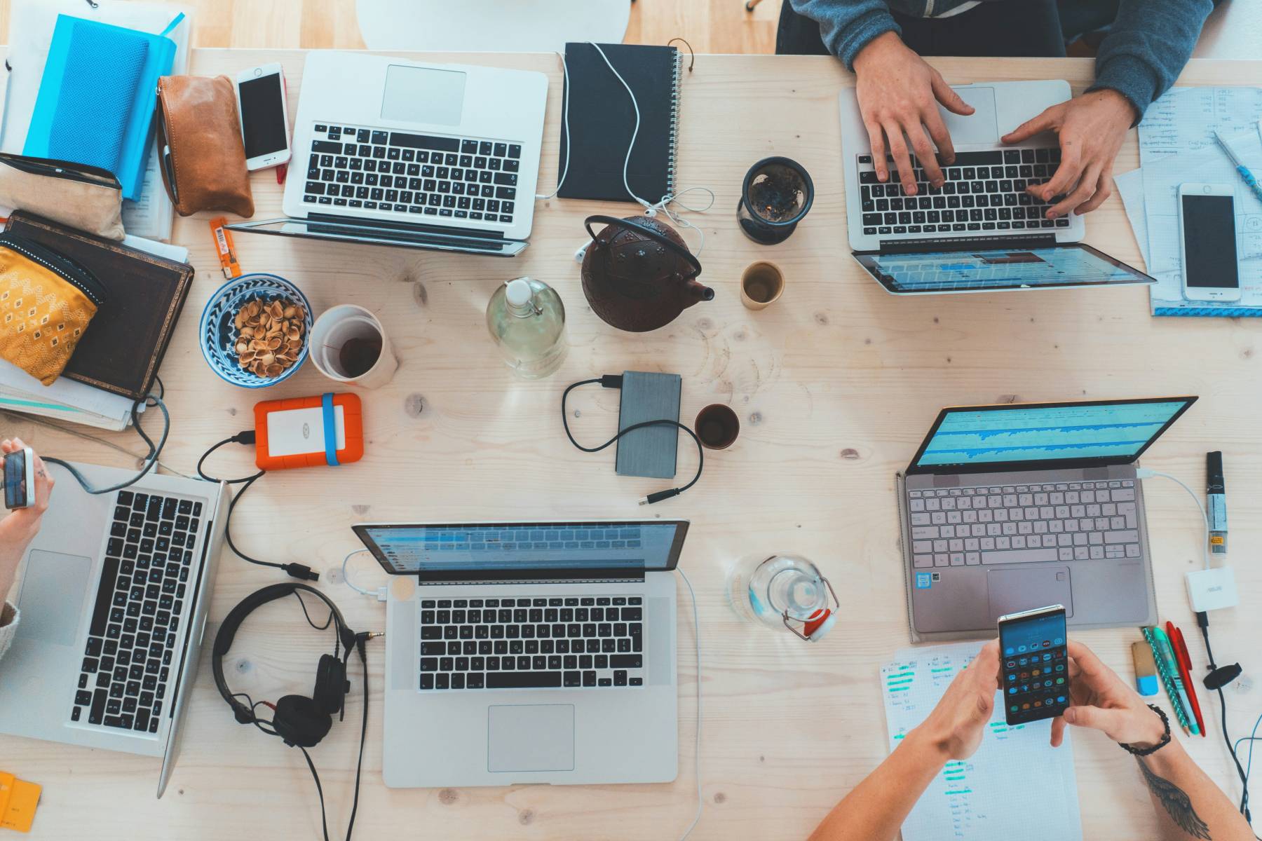 A picture of a desk with laptots, some devices and hands of two people using a phone and a laptop